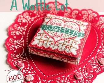 Op de afbeelding: Een rood en groen borduurwerk met de tekst "Love you a waffle lot" in de vorm van een hart. Het borduurwerk ligt op een rood kantwerk met roze hartvormige snoepjes.