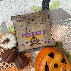 Può includere: Un cuscino ricamato a punto croce con un design autunnale che presenta una ciotola di frutta e fiori in tonalità di viola, arancione e verde. Il cuscino è su una trapunta con un design patchwork in tonalità di marrone, nero e bianco. Nell'immagine sono presenti anche due piccole zucche e una zucca intagliata.