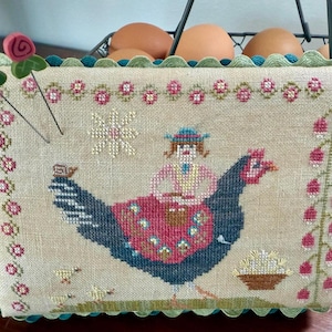 Op de afbeelding: Een kruissteek-naaldwerkontwerp van een meisje dat op een zwarte haan rijdt met een mand vol eieren. Het ontwerp is op een beige stof met een groene gegolfde rand. Het naaldwerk is ingelijst met twee spelden met rode rozenkoppen.