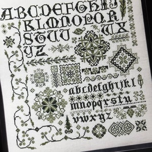 May include: Black and green cross-stitch alphabet sampler with a variety of decorative elements, including floral motifs, geometric patterns, and a feather. The sampler is framed in black and includes the text "Designed by Karen Khuba".