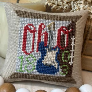 May include: A beige cross-stitch pillow with a brown border. The design features the word "Ohio" in red letters, a blue guitar, and the numbers "1803" in green. The pillow is likely handmade.