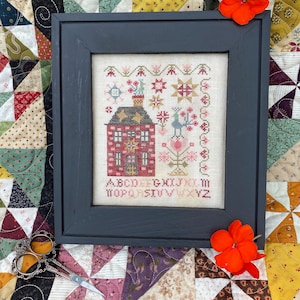 May include: A framed cross-stitch design featuring a red house with a chimney, a tree with a bird, and a floral border. The alphabet is stitched below the house. The frame is black and the background is a patchwork quilt.