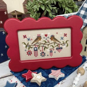 Könnte beinhalten: Ein roter Rahmen mit gewelltem Rand umrahmt eine Kreuzsticharbeit mit zwei Vögeln auf einem Ast, verziert mit patriotischen Symbolen. Das Kunstwerk zeigt amerikanische Flaggen, Sterne und Herzen in Rot, Weiß und Blau. Präsentiert auf einem blau gemusterten Stoff.