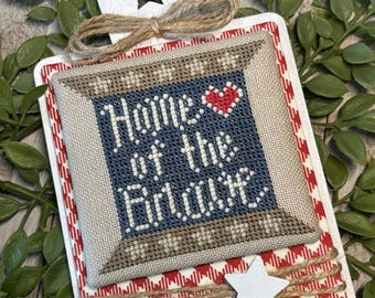 Può includere: Un design patriottico a punto croce con le parole "Home of the Brave" su sfondo blu, incorniciato da accenti beige e rossi. Il design è posto su una base a quadretti bianchi e rossi, ornato da un fiocco di spago e stelle.