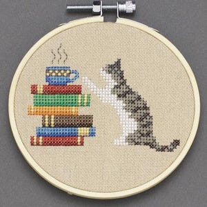 Può includere: Un cerchio da ricamo a punto croce con un gatto grigio e bianco seduto accanto a una pila di libri e una tazza di caffè blu. Il gatto sta guardando la tazza di caffè.