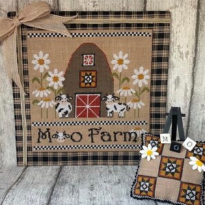 May include: A cross-stitch embroidery depicting a red barn with a white picket fence and two cows. The barn features a quilt block pattern on its side. The embroidery is framed in a black and white checked fabric with a burlap bow. The text "Moo Farm" is stitched beneath the barn.