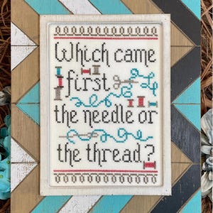 May include: Cross-stitch artwork in a white frame, with the question "Which came first the needle or the thread?" The design includes needles, thread, and spools. The frame is set within a larger, decorative wooden frame with a geometric pattern in blue, white, and brown.