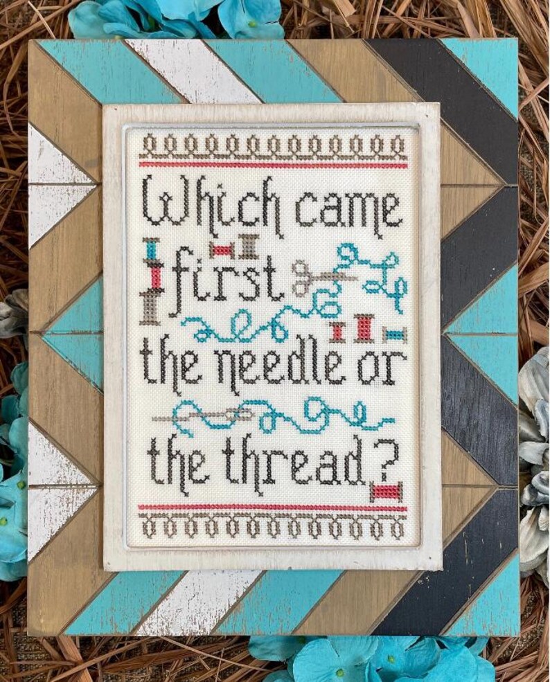 May include: Cross-stitch artwork in a white frame, with the question "Which came first the needle or the thread?" The design includes needles, thread, and spools. The frame is set within a larger, decorative wooden frame with a geometric pattern in blue, white, and brown.