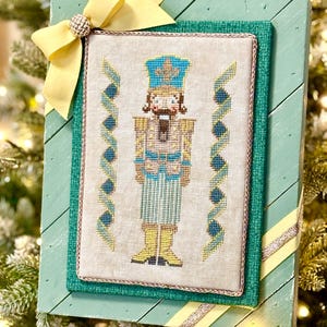 May include: A framed cross-stitch of a nutcracker soldier, with a blue and gold uniform, set against a light beige background. The frame is green with yellow and silver accents, and a yellow bow with a jeweled center adorns the top.