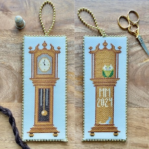 Può includere: Due ornamenti a punto croce con un orologio a pendolo dorato. Il primo ornamento ha un quadrante dell'orologio con numeri romani e il secondo ornamento ha un gufo appollaiato sulla parte superiore dell'orologio con il testo "Mimi 2023".