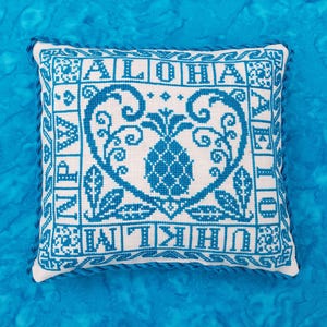 Pode incluir: Uma almofada branca com bordado azul que apresenta um desenho de abacaxi e a palavra "ALOHA" escrita em uma fonte decorativa.