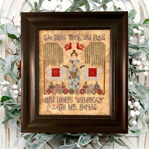May include: A framed cross-stitch sampler featuring a woman in a blue dress standing between two sheep. The text reads "She seeks wool and flax and works willingly with her hands."