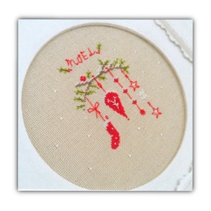 以下が含まれることがあります： ベージュの布地を背景にした白い刺繍枠。布地には、赤い糸で「Noel」の文字と、オーナメントが付いたクリスマスツリーが赤と緑の刺繍で描かれています。