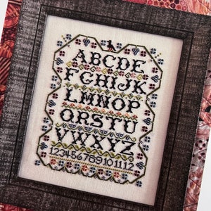 May include: A framed cross-stitch sampler featuring the alphabet, numbers 1 to 12, and floral accents in shades of red, green, blue, and yellow.