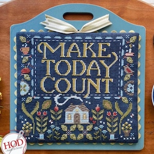 以下が含まれることがあります： 紺色の背景に金色の糸で「MAKE TODAY COUNT」と書かれたクロスステッチのサンプラー。 花柄や家のモチーフがデザインされています。 ライトブルーの木製プレートにハンドルとストライプのリボンが付いており、家の装飾に最適です。