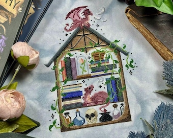May include: Cross-stitch design featuring a whimsical dragon and bookshelf scene. The design includes a dragon, books, potion bottles, and a skull. The color palette includes shades of pink, brown, green, blue, and black. The design is on a light blue background.