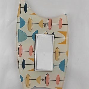 Peut inclure: Un cache-interrupteur décoratif avec un design moderne du milieu du siècle. Le couvercle présente un fond blanc avec un motif de formes géométriques colorées en bleu, jaune, rose et noir. Un interrupteur argenté et blanc est centré.