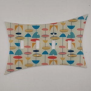 May include: A decorative light switch plate with a mid-century modern design. The plate features a repeating pattern of colorful shapes in blue, yellow, pink, and red on a cream background. The plate has four switch openings.