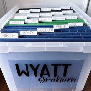 May include: A clear plastic storage box with a blue label that says "WYATT Graham". The box is filled with blue hanging file folders labeled with different stages of childhood, including "Birth", "Baby", "Toddler", "Pre-K", "Kinder", "First", "Second", "Third", "Fourth", "Fifth", "Sixth", "Seventh", "Eighth", "Ninth", "Tenth", "Eleventh", "Twelfth", "College", "Medical", and "Other".