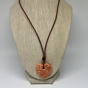 May include: A heart-shaped pendant necklace. The pendant is a light orange color with a textured surface and a hole at the top. The necklace has a brown leather cord and a cluster of beads near the pendant.