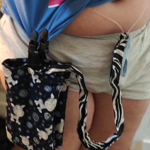 May include: A navy blue fabric pouch with a white cow pattern, attached to a black clip and a black and white zebra print strap. The pouch is hanging from a blue shirt and gray shorts.