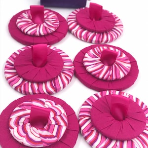 May include: Six handmade magenta fabric pin cushions with a striped center and a magenta ribbon loop. A spool of purple thread sits in the background. These sewing notions are perfect for any crafter.