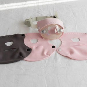 Puede incluir: Un conjunto de dispositivos médicos, que incluye una pantalla facial rosa con una cúpula de plástico transparente y una correa gris, junto con tres mascarillas en tonos rosa y gris. Las mascarillas tienen aberturas para la boca y se aseguran con broches.