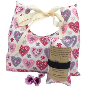 Puede incluir: Un conjunto de almohadas de confort para puerto médico con un patrón de corazones y flores en rosa, morado y blanco. Las almohadas están atadas con una cinta crema. Una almohada más pequeña está asegurada con una banda elástica negra, e incluye texto.