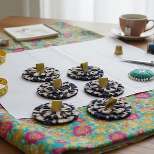 May include: A craft scene featuring sewing supplies. Six navy and cream fabric circles with gold accents are on a pattern. A yellow tape measure, scissors, and a teacup are also visible. The scene is set on a floral patterned fabric.
