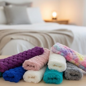 May include: A collection of colorful, plush, knitted headbands in various shades, including teal, purple, pink, and tie-dye. The headbands are arranged on a wooden surface, with a blurred bedroom scene in the background.