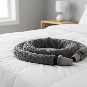 May include: A gray weighted sleep aid, coiled on a white bedspread. The cylindrical, plush item has a textured surface and a gray cuff at the end. The bed is in a room with a window and a lamp on a side table.