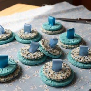 May include: Handmade fabric pin cushions in a floral pattern with a light blue ribbon accent. The pin cushions are stacked in three layers and are approximately 2 inches in diameter. The background includes scissors and pins on a floral fabric.