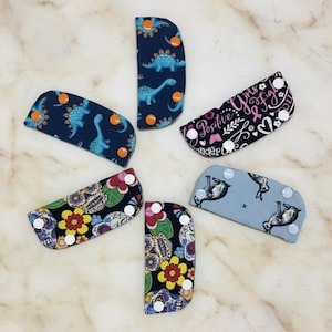 May include: A collection of fabric accessories with snap closures, featuring various patterns. Designs include dinosaurs on navy blue, sugar skulls with flowers, sloths, and a breast cancer awareness theme with the text "Positive Vibes Fight".