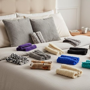 May include: Various coloured, rolled-up blankets with a plush, furry texture are displayed on a bed. Colours include purple, blue, black, and cream. The scene is set with pillows and a neutral-toned bedspread. The blankets appear soft.