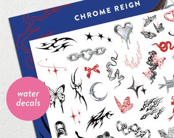 Puede incluir: Una hoja de calcomanías de agua con varios diseños, incluyendo mariposas, cadenas, estrellas y patrones tribales. Las calcomanías son en negro, plata y rojo, con las palabras "water decals" en un círculo rosa. La hoja está etiquetada como "CHROME REIGN".