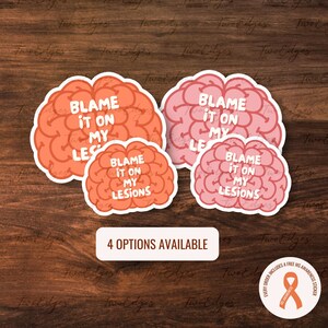 Pode incluir: Quatro opções de adesivos com o contorno de um cérebro com o texto "Blame it on my lesions". Os adesivos são em tons de rosa e laranja. O texto é branco. Os adesivos estão sobre um fundo de madeira.
