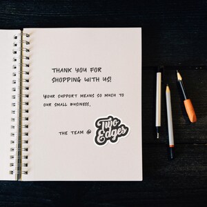 May include: A handwritten thank you note on a white spiral notebook with the text "THANK YOU FOR SHOPPING WITH US! YOUR SUPPORT MEANS SO MUCH TO OUR SMALL BUSINESS. THE TEAM @ Two Edges".
