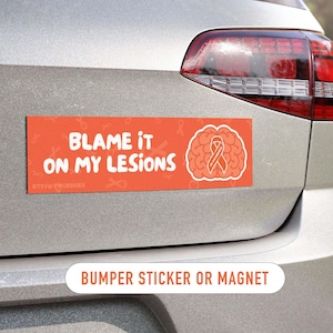 May include: A rectangular orange bumper sticker or magnet featuring the white text "BLAME IT ON MY LESIONS" and a white outline of a brain containing an awareness ribbon. The background has a subtle pattern of small, faint orange awareness ribbons.
