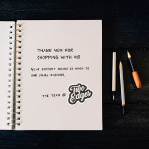 May include: A handwritten thank you note on a white spiral notebook with the text "THANK YOU FOR SHOPPING WITH US! YOUR SUPPORT MEANS SO MUCH TO OUR SMALL BUSINESS. THE TEAM @ Two Edges".
