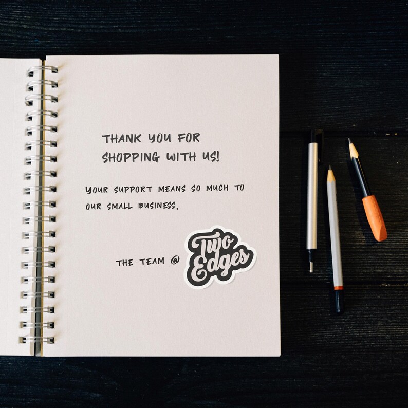 May include: A handwritten thank you note on a white spiral notebook with the text "THANK YOU FOR SHOPPING WITH US! YOUR SUPPORT MEANS SO MUCH TO OUR SMALL BUSINESS. THE TEAM @ Two Edges".