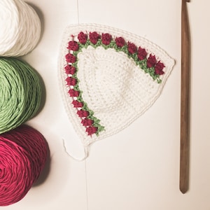 May include: A white crocheted piece with a red rose and green leaf border, alongside white, green, and red yarn balls, and a wooden crochet hook. The handmade item is likely part of a larger craft project.
