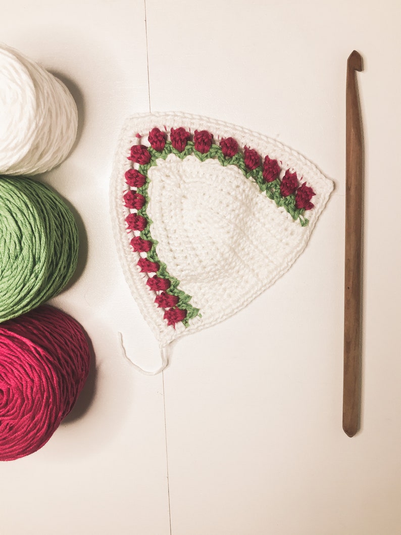 May include: A white crocheted piece with a red rose and green leaf border, alongside white, green, and red yarn balls, and a wooden crochet hook. The handmade item is likely part of a larger craft project.