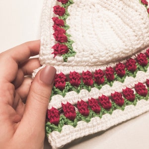 May include: A white crochet top with red rose and green leaf detailing. The top features a scalloped edge and is held by a hand. The crochet work is visible, showcasing the texture and craftsmanship of the handmade garment.