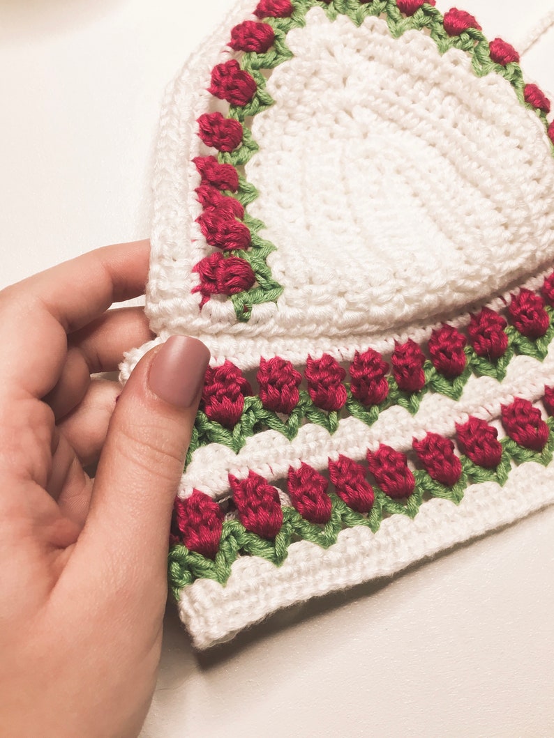 May include: A white crochet top with red rose and green leaf detailing. The top features a scalloped edge and is held by a hand. The crochet work is visible, showcasing the texture and craftsmanship of the handmade garment.