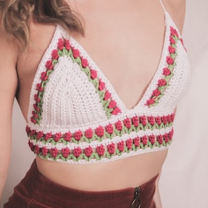 May include: White crochet bralette with red and green floral detailing. The top features a triangle shape with a halter neck and a band of flowers along the bottom edge. The floral pattern is repeated along the neckline.
