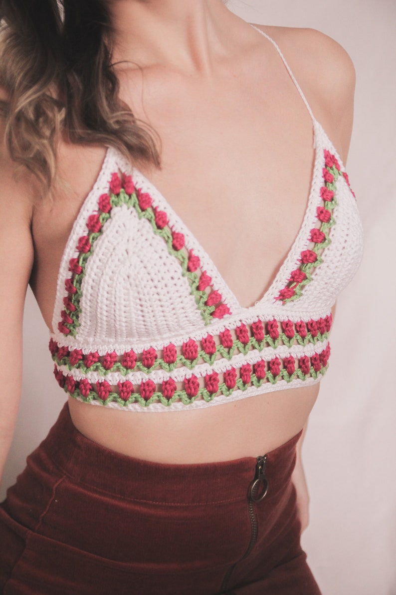 May include: White crochet bralette with red and green floral detailing. The top features a triangle shape with a halter neck and a band of flowers along the bottom edge. The floral pattern is repeated along the neckline.