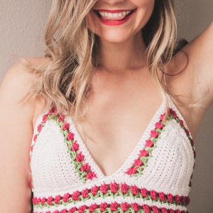 May include: A white crochet top with a V-neck design. The top is adorned with a pattern of red flowers and green leaves along the edges. The top is a handmade item.