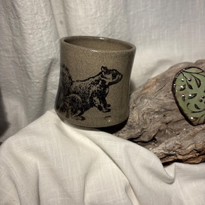 Könnte beinhalten: Eine handgefertigte Keramiktasse mit einem beigefarbenen, gesprenkelten Finish und einer schwarzen Eichhörnchen-Illustration. Die Tasse ist zylindrisch und scheint aus Steinzeug zu sein. Ein kleines grünes Dekorationselement mit einem Blattmuster ist im Hintergrund zu sehen.