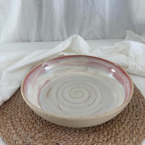 Könnte beinhalten: Eine runde Keramikschale mit gesprenkelter beiger Außenseite und einer glatten Innenseite mit Spiralmuster am Boden. Der Rand ist in einem sanften Rosaton glasiert, der in eine cremeweiße Mitte übergeht und einen subtilen Verlauf erzeugt.