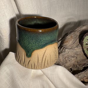 Könnte beinhalten: Eine handgefertigte Keramik-Tasse mit einem einzigartigen Design. Die obere Hälfte ist in einem tiefen Grün glasiert, das in einen beigefarbenen, strukturierten Sockel übergeht. Der Rand ist dunkelbraun. Die Tasse hat eine zylindrische Form und ist für heiße oder kalte Getränke geeignet.